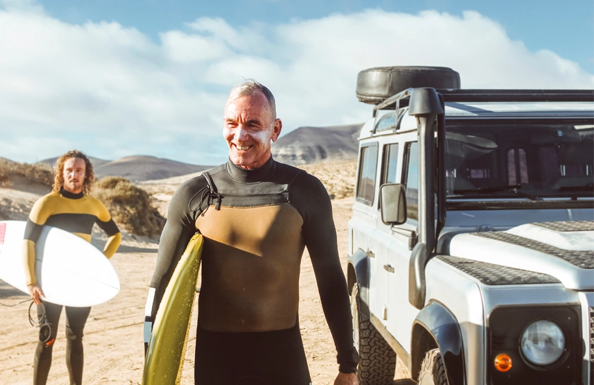 senior male on the beach ready to surf with a younger man behind him. Both are wearing sunscreen and they are walking past a Jeep.